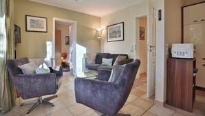 A living room with two armchairs, a sofa, and a glass coffee table. There is a kitchen area with a microwave on a wooden cabinet. - Moderne Strandvilla Appartement 2 (Timmendorfer Strand)