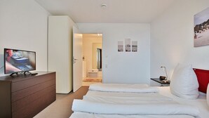 A hotel room with a bed, a TV on a dresser, a desk with a lamp, and a door leading to another room. - Villa Mér Baltique Appartement 13 (Timmendorfer Strand)