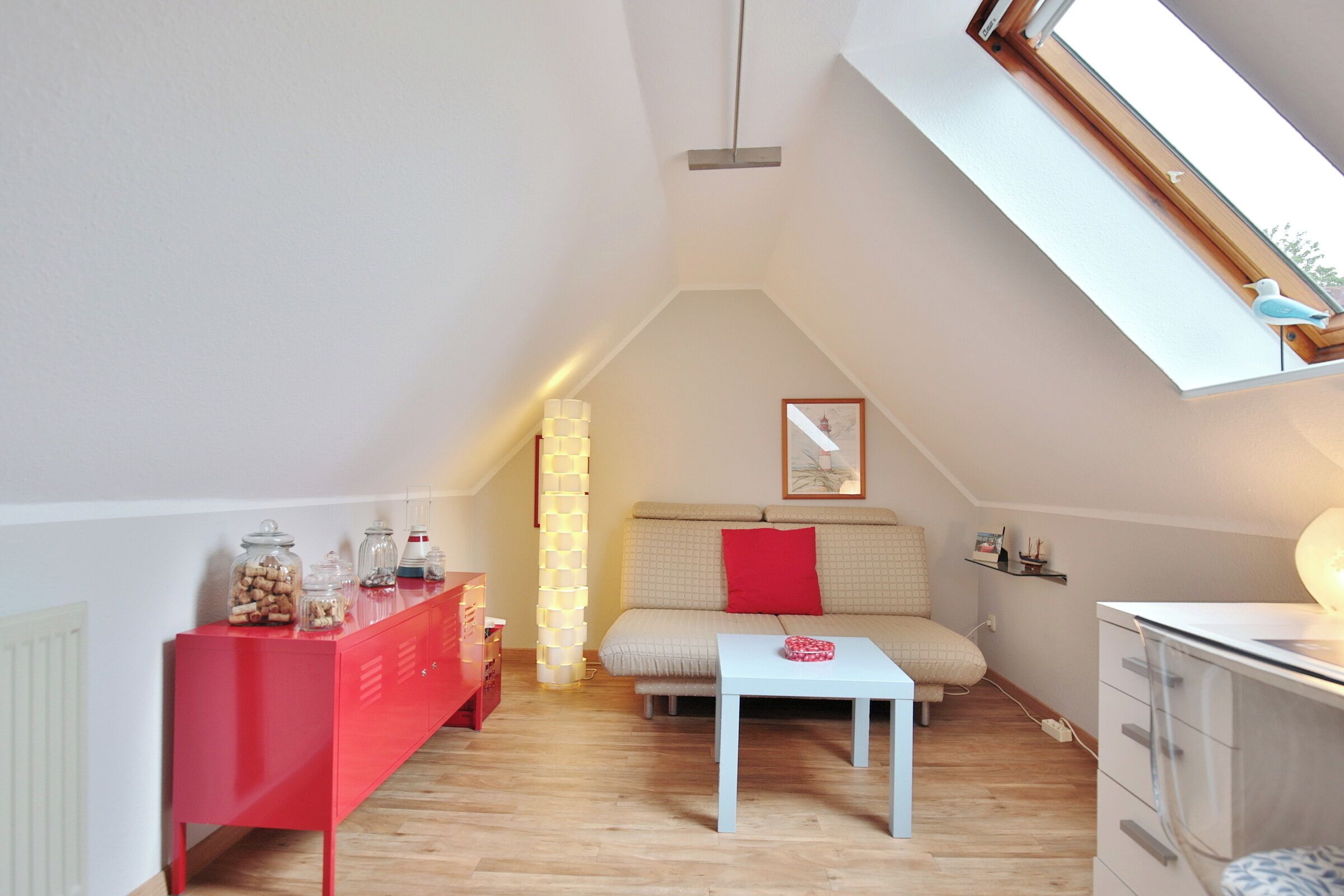 Een knus loft slaapkamer met een schuine plafond, een rode dressoir, een witte tafel, een beige bank en een dakraam.