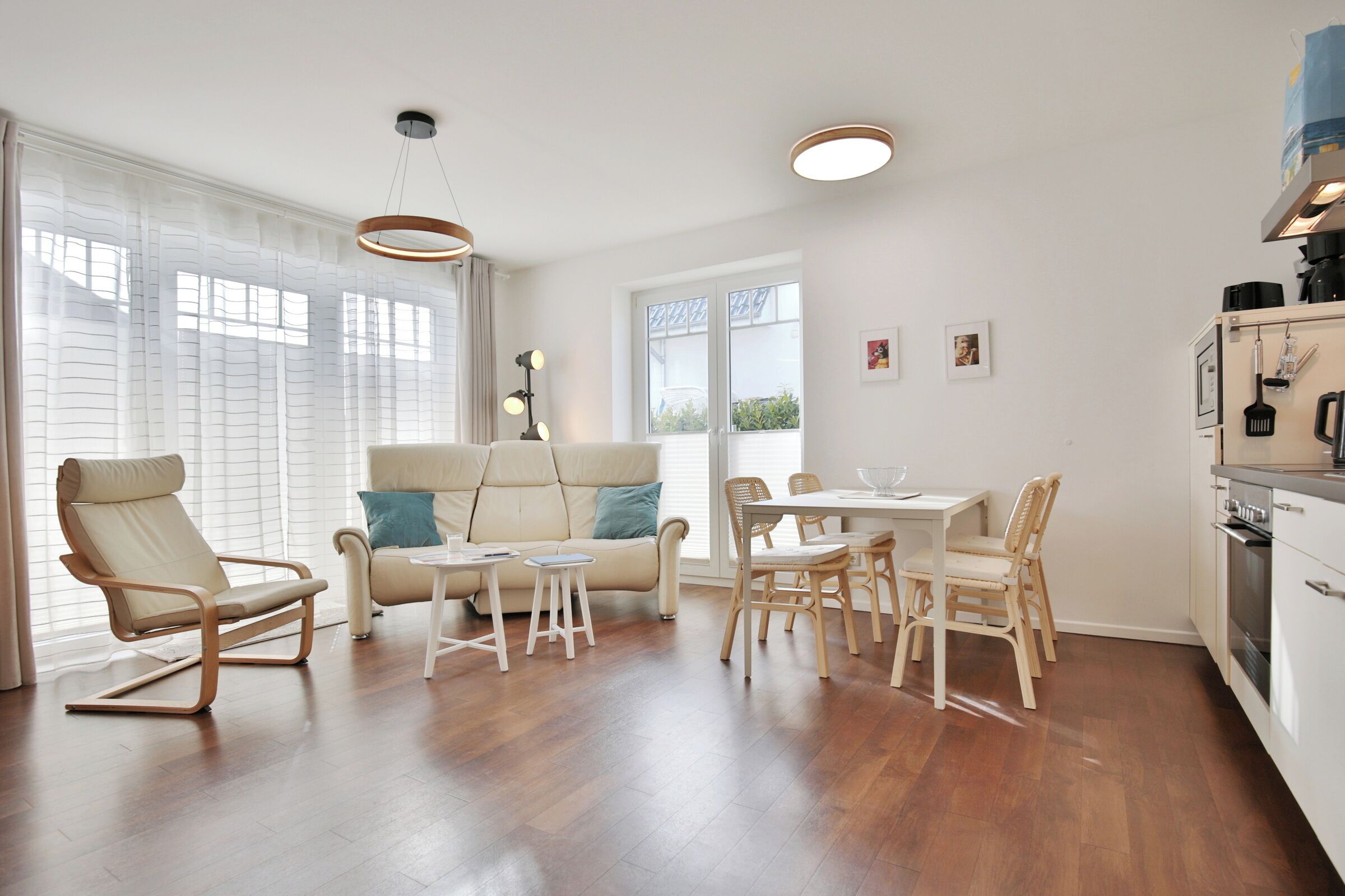 Een moderne woonkamer met een bank, een eettafel met stoelen, een keuken met inbouwapparatuur en grote ramen met dunne gordijnen.