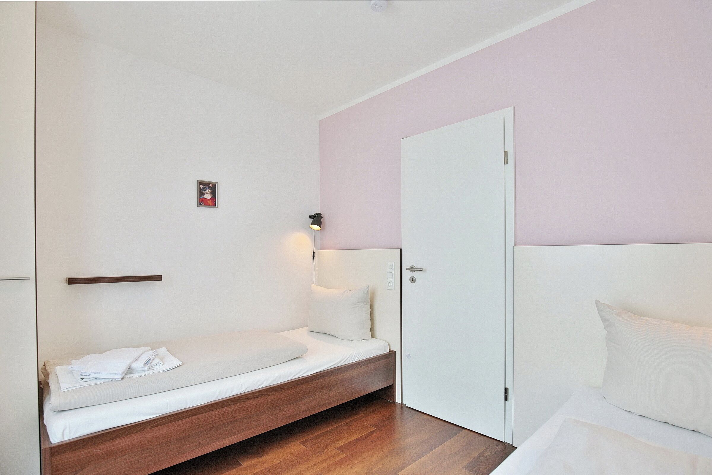 Una cama individual con estructura de madera y ropa de cama blanca, una mesita de noche con una lámpara, y una puerta en una pared rosa.