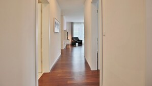 Un corridoio con pavimento in legno, pareti bianche e una lampada a soffitto. Si nota una porta a destra e una sedia in lontananza.