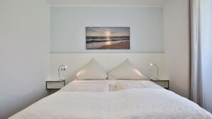 Una habitación de hotel moderna con una cama grande, dos mesitas de noche y un televisor de pared mostrando una escena de playa.
