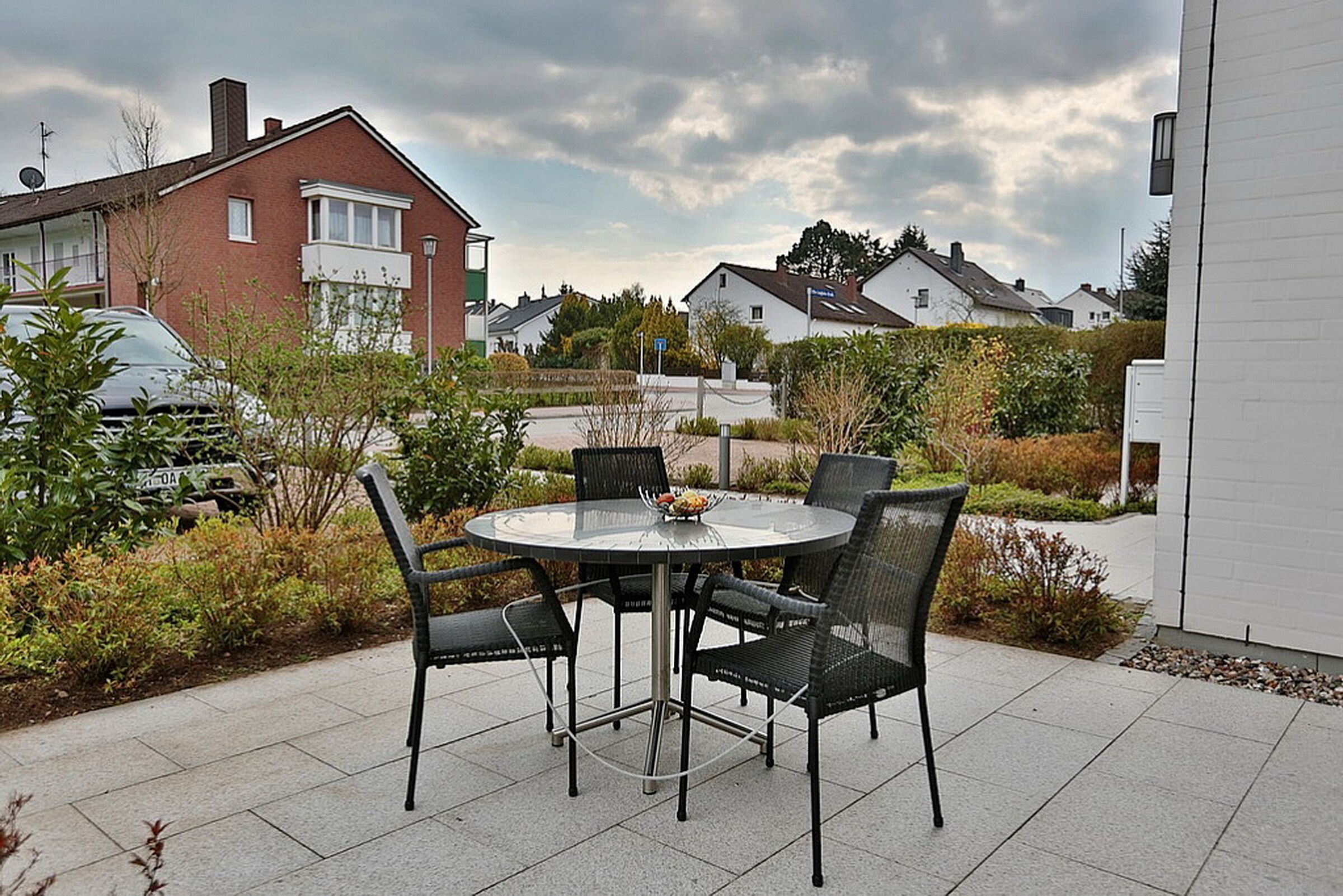 A patio with a round table and four chairs, surrounded by a garden with various plants and a white building in the background.