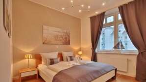 Un dormitorio con cama, lámparas de noche, ventana con cortinas, radiador y una obra de arte colgada en la pared.