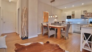 Una cocina moderna con armarios blancos, una zona de comedor con mesa y sillas de madera, y una lámpara colgante.