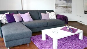 Moderni olohuone, jossa on harmaa kulmasohva, valkoinen sohvapöytä ja violetti, pörröinen matto.