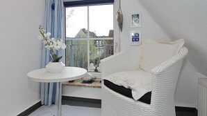 En hvid fletstol med pude, et lille hvidt bord med en vase fyldt med hvide blomster, og et vindue med blå gardiner.