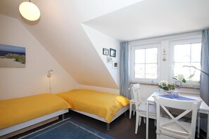 Une petite pièce lumineuse, avec un lit, une table entourée de chaises, une fenêtre avec des rideaux et un luminaire fixé au mur.