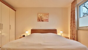 A neatly made bed in a hotel room with a wooden headboard, two bedside lamps, a wall-mounted artwork, and a window with curtains. - Villa Mér Baltique Appartement 20 (Timmendorfer Strand)