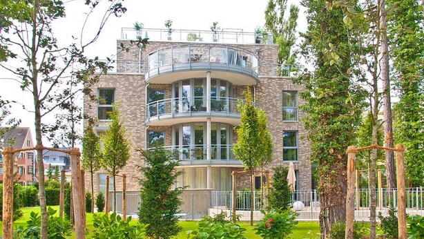 Een meerlaags woongebouw met een balkon op de bovenste verdieping, omgeven door groen en bomen.