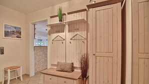 Une pièce comprenant un banc en bois, un petit tabouret, une étagère fixée au mur avec une plante et un vase contenant des plantes séchées.