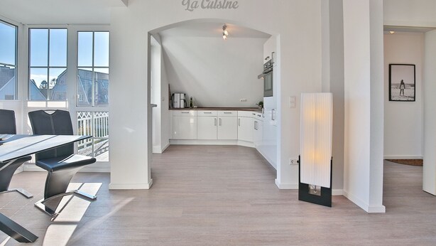 Eine moderne Küche mit weißen Schränken, ein Essbereich mit Tisch und Stühlen und ein Ausblick ins Freie durch große Fenster.