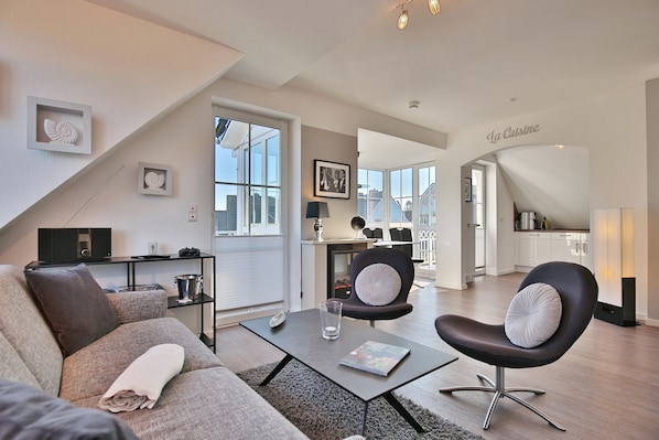 Ein modernes Wohnzimmer mit Sofa, Couchtisch und Sessel. Im Hintergrund ist eine Küche zu sehen.