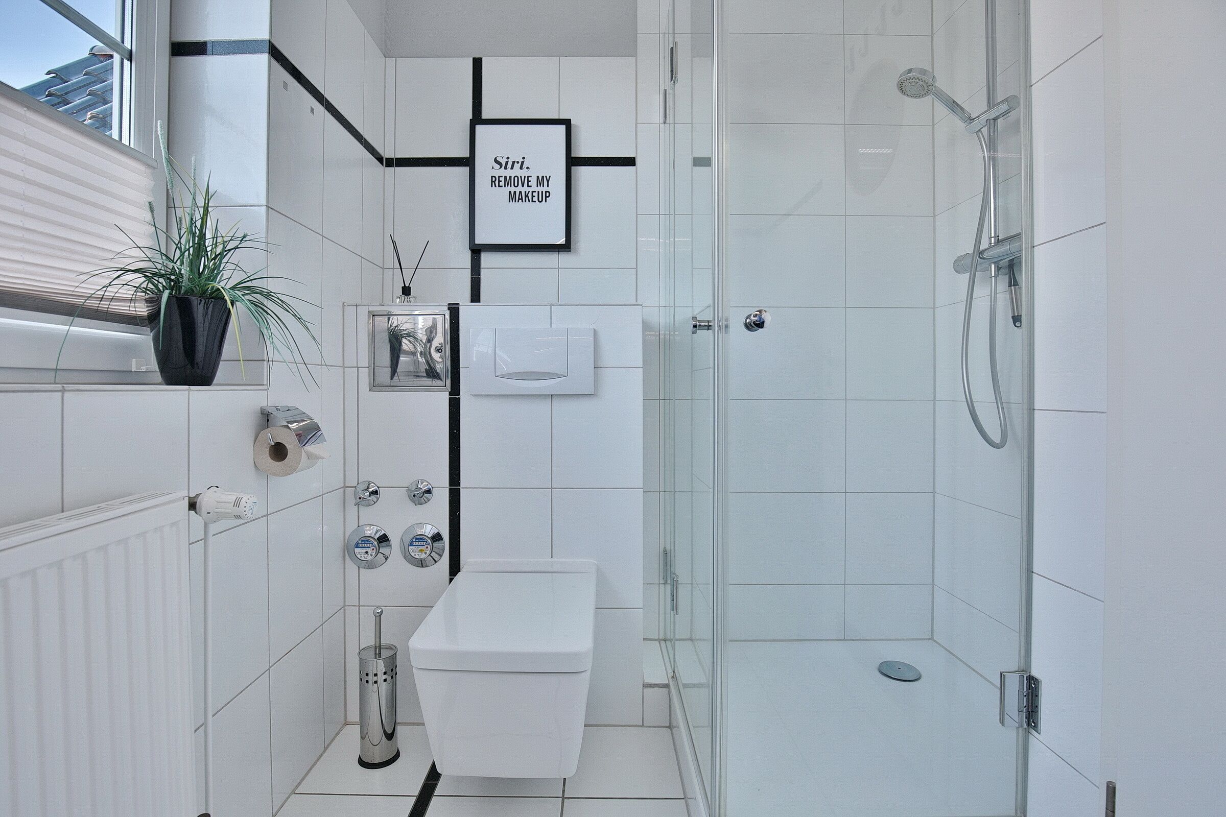 Un baño moderno con inodoro, bidé y ducha. Hay un cartel enmarcado en la pared y una planta en el alféizar.