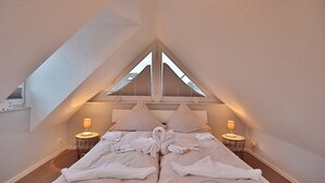 Ein Schlafzimmer mit schrägen Wänden, zwei Nachttischen mit Lampen, einem Bett mit Handtüchern und einem Fenster mit Jalousien.