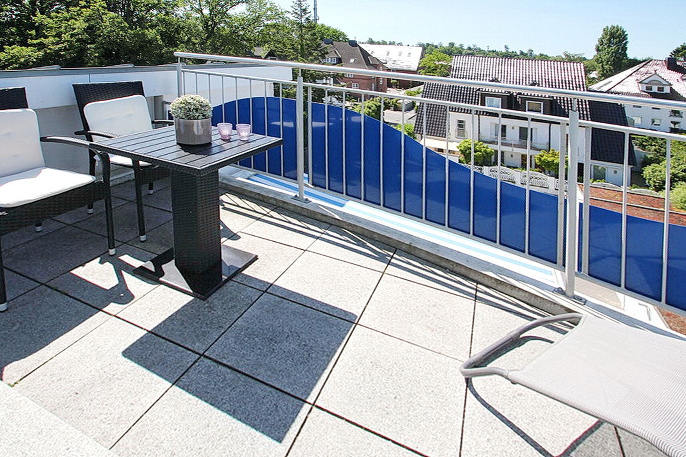 A balcony with a table and chairs, a potted plant, and a view of a blue fence and buildings.