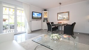 Ein modernes Wohnzimmer mit Glastisch, Esstisch und Stühlen. Ein Flachbildfernseher ist an der Wand montiert, sowie ein Schrank und ein Spiegel.