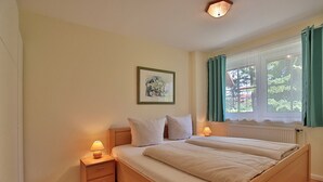 A double bed with a wooden headboard, two bedside tables with lamps, a framed picture on the wall, and a window with curtains. - Gorch-Fock-Park 57 Apartment 17 Beach pleasure (Timmendorfer Strand)