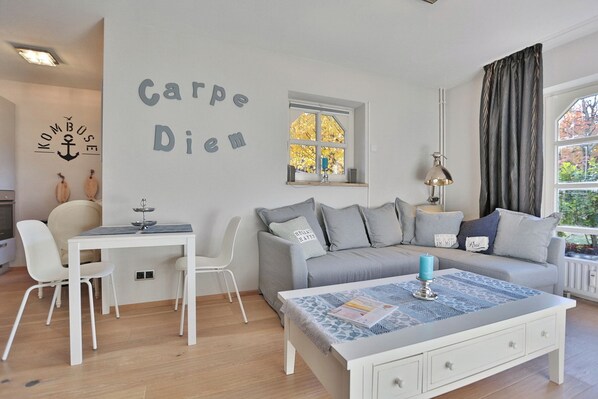 Una sala de estar moderna con un sofá blanco, cojines grises, una mesa de centro blanca con una vela azul y una ventana con cortinas.