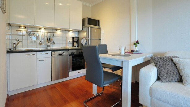 Una cocina compacta con armarios blancos, electrodomésticos de acero inoxidable, una mesa de comedor con sillas y un sofá con un cojín.