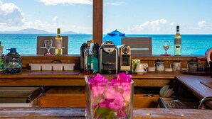 A wooden bar counter with various bottles and glasses, a coffee maker, and a view of the ocean. - Conch Beach Cabins - Rumbus Beach (Johnson's Point)