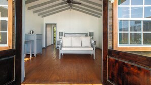 A bedroom with a wooden floor, a bed with white linens, a ceiling fan, and a window with a view of the outdoors. - Conch Beach Cabins - Rumbus Beach (Johnson's Point)