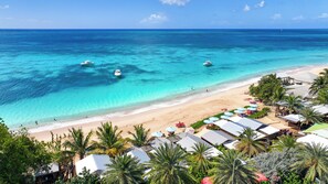 A beach with clear turquoise waters, a sandy shore, and several boats. There are umbrellas and beach chairs, and a building with a sloped roof. - Conch Beach Cabins - Rumbus Beach (Johnson's Point)