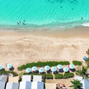 An aerial view of a beach with clear turquoise water, a sandy shore, and rows of beach umbrellas and lounge chairs.