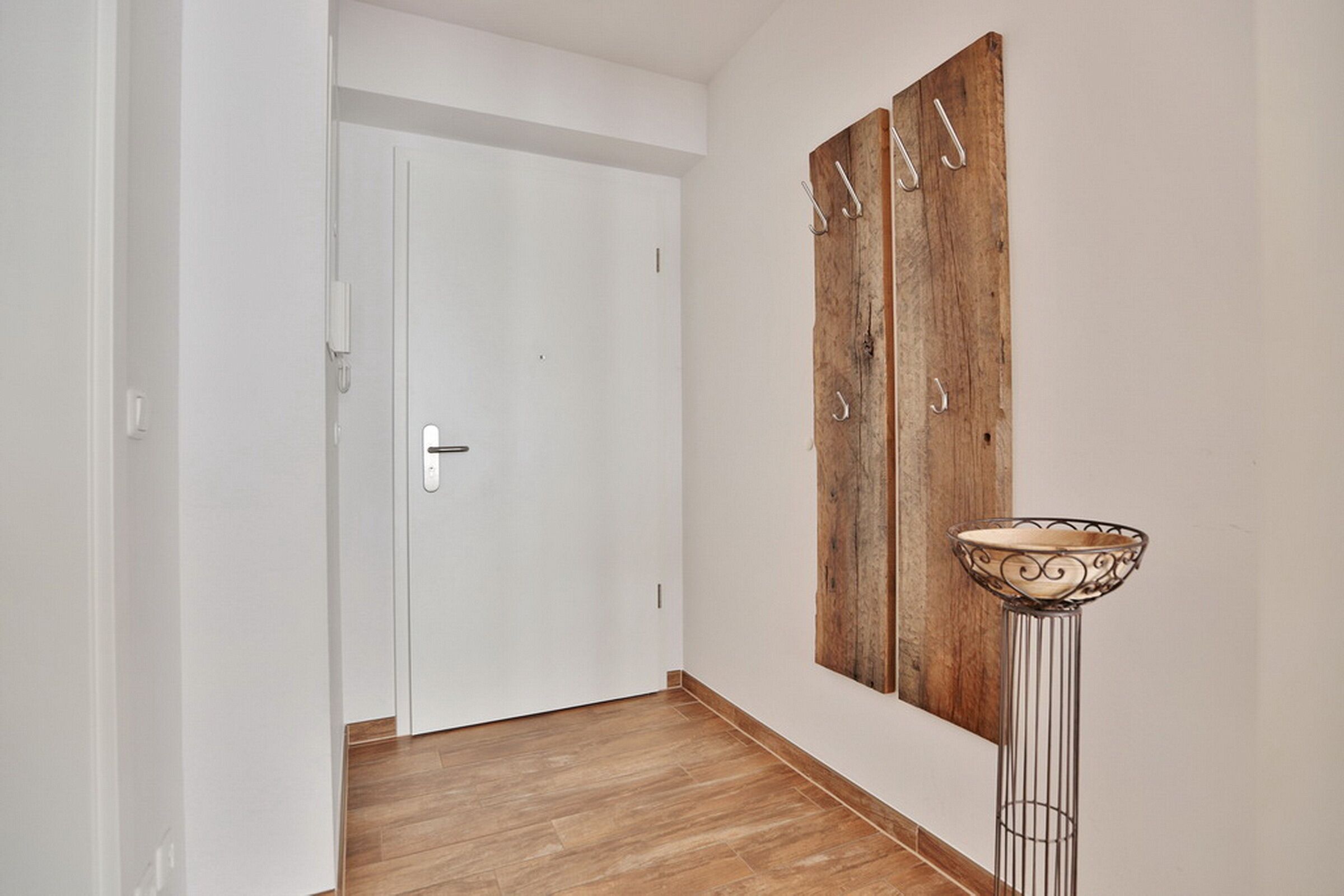 Un pasillo con una puerta blanca, un muro de madera con ganchos metálicos y una lámpara de pie.