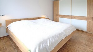 A bedroom with a wooden bed frame, white bedding, a nightstand with a lamp, and a large wardrobe with a wooden panel design. - Die Wolburg Appartement 6 (Timmendorfer Strand)