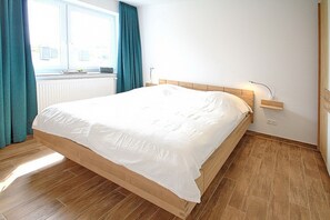 Un lit bien fait dans une pièce au plancher de bois, avec une fenêtre ornée de rideaux et une table de chevet sur laquelle se trouve une lampe.