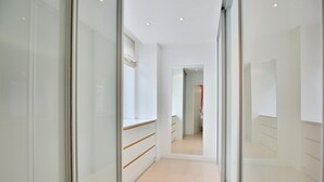 Un couloir étroit aux murs blancs et au sol carrelé, qui mène à une pièce dotée d’un miroir et d’une porte.