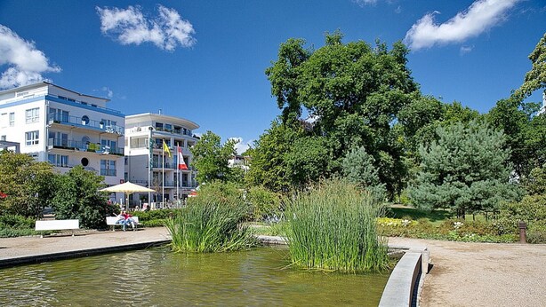Un parc paisible avec un étang, des bancs et un bâtiment à plusieurs étages doté de balcons.