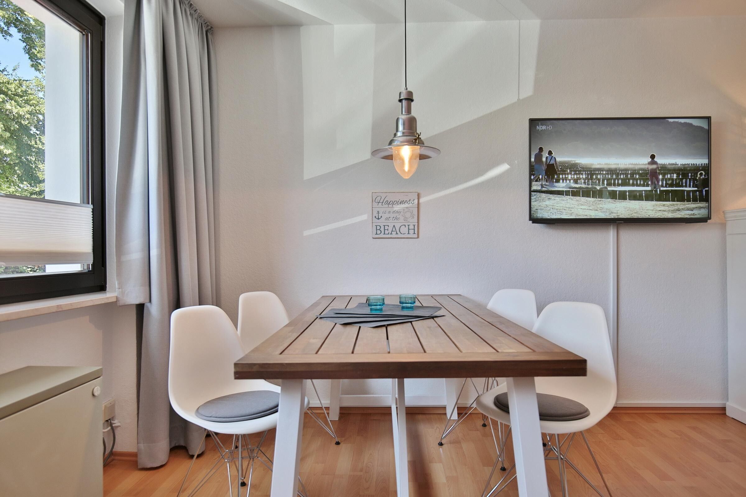 A dining area with a wooden table, white chairs, a hanging light fixture, a television mounted on the wall, and a window with curtains.