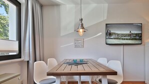 A dining area with a wooden table, white chairs, a hanging light fixture, a television mounted on the wall, and a window with curtains. - Strandkoje Kastanienallee (Timmendorfer Strand)