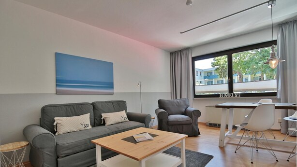 A modern living room with a grey sofa, a wooden coffee table, a grey armchair, and a dining table with chairs. A large window provides a view of buildings outside. - Strandkoje Kastanienallee (Timmendorfer Strand)