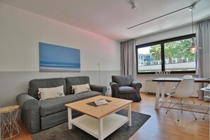 A modern living room with a grey sofa, a wooden coffee table, a grey armchair, and a dining table with chairs. A large window provides a view of buildings outside.