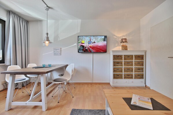 A modern room with a wooden table, white chairs, a television mounted on the wall, and a cabinet with wooden drawers.