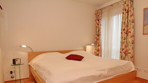 Quarto com cama de madeira, roupa de cama branca, almofada vermelha, mesa de cabeceira com abajur e cortina floral.