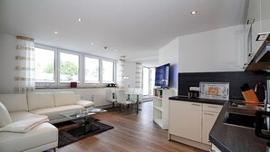 Ein modernes Wohnzimmer mit Sofa, Couchtisch und Fernseher. Es gibt eine Küchenzeile mit Kaffeemaschine und einem Schild, das Kaffee- und Cappuccino-Optionen anzeigt.