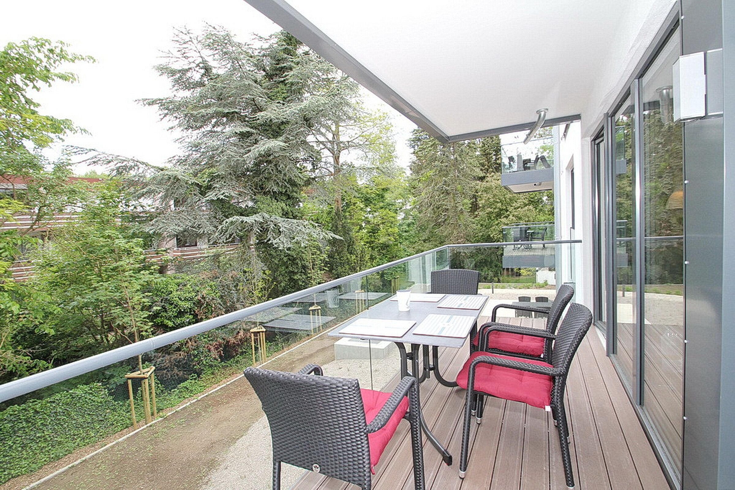 A balcony with a glass railing, outdoor furniture, and a view of the surrounding greenery and trees.