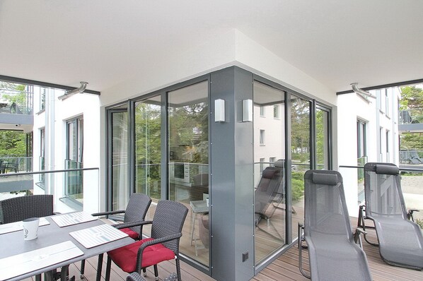 A modern balcony with glass walls, wicker chairs, and a table with a red cushion. - Villa Stern Appartement 7 (Timmendorfer Strand)