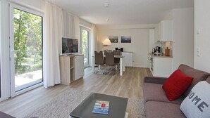 A modern living room with a sofa, a coffee table, and a TV. There is a dining area with a table and chairs, and a kitchen area with cabinets and a countertop. - Villa Stern Appartement 9 (Timmendorfer Strand)