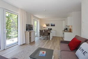 Uma sala de estar moderna com um sofá, uma mesa de centro e uma televisão. Há uma área de jantar com uma mesa e cadeiras, e uma cozinha com armários e bancada.