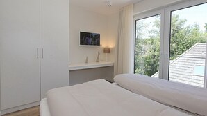A bedroom with a bed, a TV mounted on the wall, a bedside table with a lamp, and a window with curtains. - Villa Stern Appartement 9 (Timmendorfer Strand)