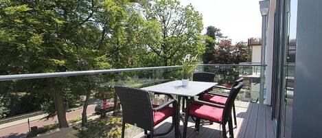 Un balcon avec une table et des chaises, donnant sur une rue bordée d’arbres et où est garée une voiture.