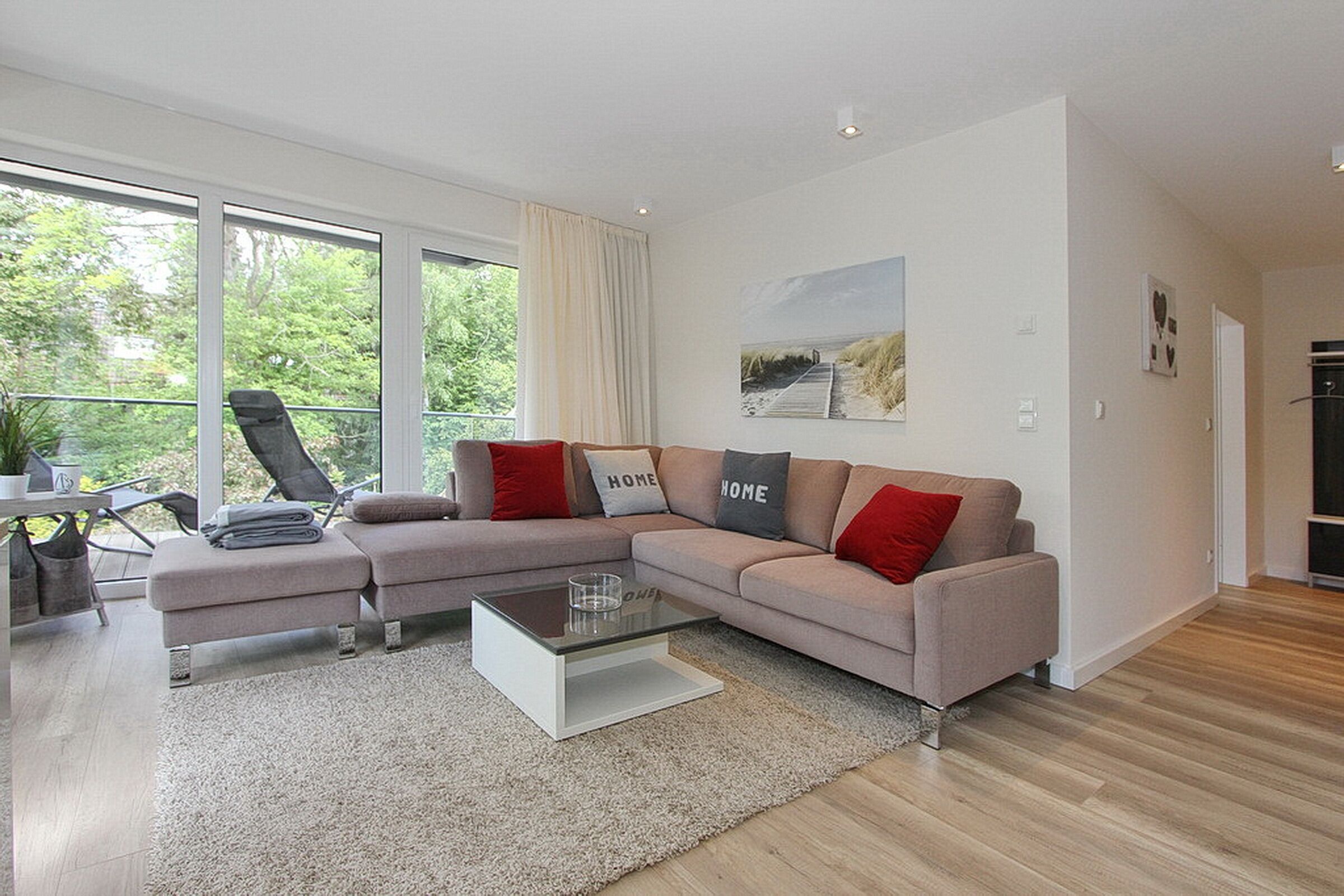 Een moderne woonkamer met een beige modulaire bank, een glazen salontafel en uitzicht op groen door grote ramen.