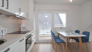 A modern kitchen with white cabinets, a built-in oven, and a coffee maker. There is a dining area with a table and blue chairs. - Ferienanlage Bergstrasse 6 Appartement 7 (Timmendorfer Strand)