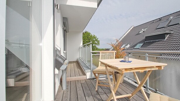 A balcony with a wooden table and chairs, a glass door, and a view of neighboring buildings. - Ferienanlage Bergstrasse 6 Appartement 7 (Timmendorfer Strand)